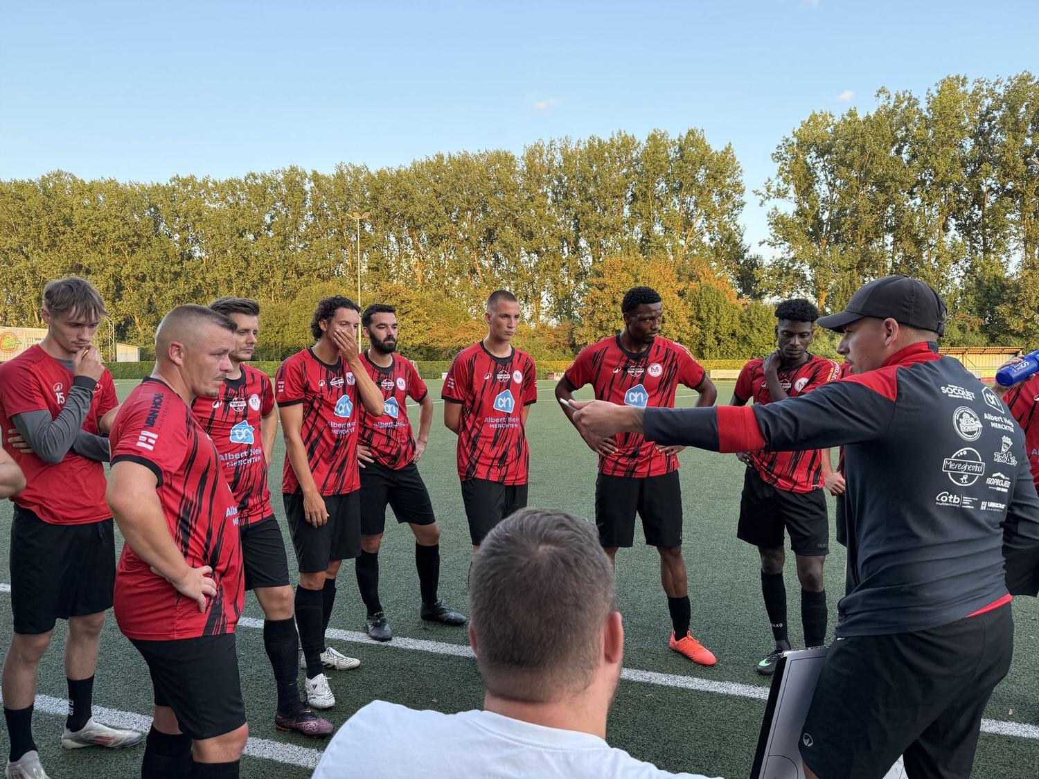 De trainer geeft instructies aan de spelers in wedstrijdtenue langs het veld.
